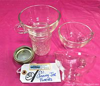 Three clear glass wide mouth canning funnels with handles shown from angled front view alongside a jar lid and handwritten label.