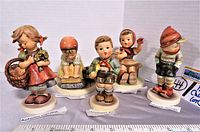 Front view of the five Hummel Goebel figurines with labels displayed in front of each figure on white bases.