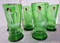 Front view of five emerald green Mary Gregory tumblers with Victorian children painted on the glass, placed upright next to a ruler showing height.