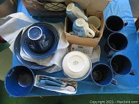 Full view of both blue stoneware dish sets including stacked plates, bowls, mugs, and boxed flatware with blue handles on blue tablecloth.