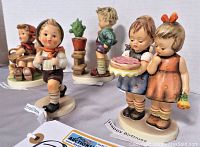 Five Goebel Hummel figurines displayed on table with close-up view showing details and tags identifying figurines by name.