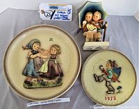 Lot showing three vintage Goebel Hummel items arranged on a surface: two plates and one figurine. The plates have distinct images with years and numbers visible. Figurine shows two children under umbrella.