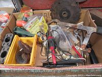 Photo showing a box with miscellaneous hand tools like wire cutters, saw blades, brushes, and small hardware items in a yellow plastic bin