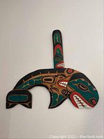 Wood carved indigenous whale wall hanging painted in black, green, and red with traditional motifs, displayed on a white wall.