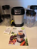 Full view of blender base, several blending cups with lids, blade assembly, and instruction manual.