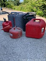 Fuel cans including two grey metal cans, three red plastic cans with spouts and one red metal gasoline can with attached funnel, all placed on concrete outside.