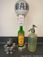 Full view of the Cutty Sark bottle lamp with white bar globe, pewter drinking set, and soda siphon bottle on countertop.