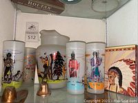 Photo showing ceramic pitcher, two glasses with Oklahoma Indians designs, small copper bell, small cowboy hat, and decorative box with Native American chief image.
