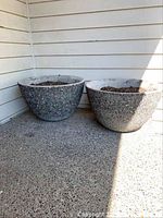 Two large round concrete planters with exposed aggregate finish sitting on a concrete floor against white siding wall.