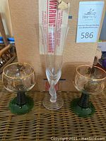 Two glasses displayed, showing the green twisted stems and gold floral design with a champagne flute in the background.