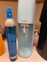 Light blue SodaStream carbonator with one new sealed CO2 canister standing beside it on a kitchen counter.