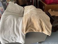Photo showing two blankets draped over a table, one off-white and one beige, both plush and lightweight.