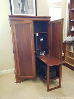 Wooden foldable desk cabinet with doors open and fold-out surface extended, shown empty in a room.
