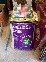 Box of Buffalo Snow artificial snow with assorted decorative fabrics on top, including checkered and gold fabric pieces.