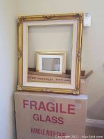 Seven large and one small gold decorative picture frames stacked against a wall beside fragile glass packing boxes.