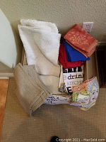 Photo showing assorted folded linens including white towel, red and blue cloths, beige small rug, and printed kitchen towel with letters.