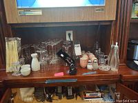 Wide shot of assorted barware items, stemware glasses, ceramic sake set, crystal vases, dispensers, and small accessories arranged on a wooden surface.