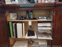 Open wooden cabinet displaying contents including two cordless landline phones with bases, multiple hands-free headsets, binders, notebooks, plastic organizers filled with papers, a black printer stand, and assorted stationary items.