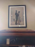 Framed black and white image showing two dancing figures with a third figure in the background, mounted on a wall above a wooden piece of furniture.