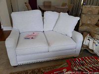 Front view of light linen loveseat with loose back pillows and brass nailhead trim, placed on carpet near dining area