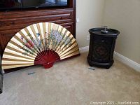 Photo showing large gold folding wall fan with floral design, measuring approximately 35x61 inches, placed on floor beside the Bombay chest