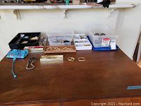 Wide photo showing multiple clear plastic storage bins filled with various costume jewelry items, plus some loose pieces on the table including a strand of pearls, bracelets, and boxes.