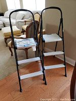 Two black steel frame step ladders with three aluminum steps each, positioned on wooden floor inside a living area.