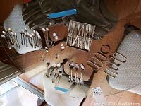 Wide view of assorted silverware including forks, spoons, knives of different patterns laid on chairs and table.