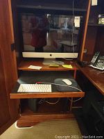 Wooden hutch shelf holding silver aluminum Apple iMac with white wired keyboard on slide-out tray, white Magic Mouse on gray pad, Apple Watch charging puck beside iMac, black tower device below.