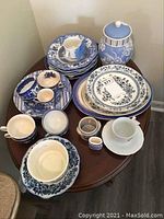 Full overview of the blue transferware collection on a round table showing plates, cups, bowls, jar, and small items in blue and white patterns.