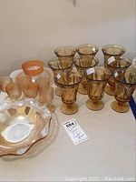 Full view of peach amber carnival glass bowl, six small glasses, Little Herbert cup, three plates, and nine dark amber stemmed glasses on table.