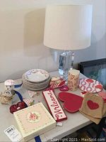 Six floral patterned plates stacked with a matching bowl, floral box with napkins, heart-shaped hot plate, wooden 'Welcome' sign, heart dishes, votive candle, heart ornaments, heart burlap bags, red glass heart and ceramic poodle night light. Clear glass lamp with white shade behind items.