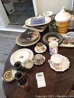 Photo showing an assortment of ceramic plates, teacups with saucers, a ceramic cookie jar shaped like an ice cream cone, candleholders, a ceramic owl figurine, mugs, and platters arranged on a dark wood surface.