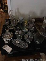 Clear glass lot on black surface with visible glasses, jars, decanter, custard cups, dishes, candlesticks, votives and cone-shaped glass item.