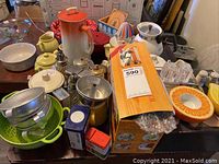 Photo showing various vintage kitchenware items including Corning glass teapot, metal mugs, bright green strainer, cookie cutters, molds, and orange ceramic dish.
