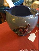 Blue ceramic pitcher with grape and leaf relief decoration on a red tablecloth, shown from front angle.