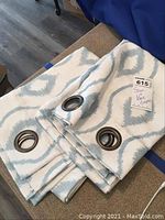 Pair of folded linen cafe pole drapes in white with pale blue geometric diamond pattern and large metal rings for hanging.