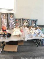 Full view of table with 25 dolls displayed, including boxed Brinn dolls and loose Dolls of the Nations and other porcelain/fabric dolls in various costumes and hairstyles.