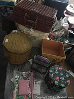 Photo showing large brown wicker picnic basket on top, 8-sided woven metal tin, poppy patterned box without lid, black cardboard box, clear and pink purses, butterfly patterned hexagonal tin box