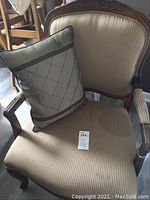 Photo of upholstered armchair showing striped upholstery with carved wooden frame and pillow on seat, including item tag visible on seat.