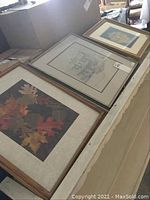 Three framed art pieces laid on table: fall leaves print, cottage pen and ink drawing, and Needlepoint US Naval Academy artwork grouped side by side.