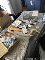 Photo showing box of linens including crocheted mantle scarf, doilies, curtain fabric, baby hat, napkins, whisk broom, and small miscellaneous items.