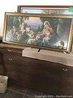 Full view of large framed vintage wall art depicting the Blessed Mother with Christ and angels, leaning on furniture.