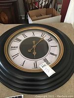 Front view of large round wall clock showing Roman numerals and detailed hour and minute hands. Clock has black frame and gold inner rim.