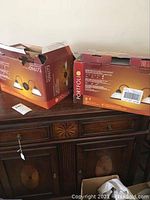 Two boxes of Portfolio dual light sconces on wooden sideboard, both boxes open showing the same product image.