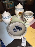 Three floral canisters with blue geometric band near the top, one lid missing, arranged around five stacked plates with blue floral design.
