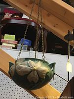 Green ceramic Roseville hanging pot with clematis flower design, suspended by metal chain, against a background of wooden beams and stacked boxes