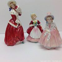 Three Royal Doulton figurines, Valerie, Christmas Morn, and Lily displayed together showing their sizes and colors.