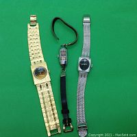 Photo showing three ladies wristwatches laid out on a green surface: a gold-tone Wittnauer with wide link bracelet, a small Oplex watch with black leather strap, and a silver Wittnauer watch with mesh bracelet.