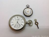Two pocket watches laid flat alongside a wind key: larger Elgin with white face, Roman numerals, and 'Elgin Natl Watch Co.' markings; smaller simpler Spiegelhalter watch